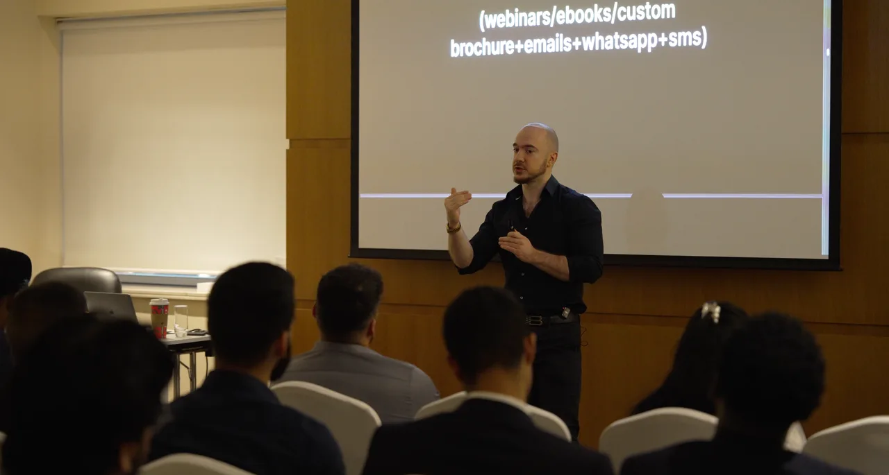 Mikail speaking and gesturing in front of a seated audience.